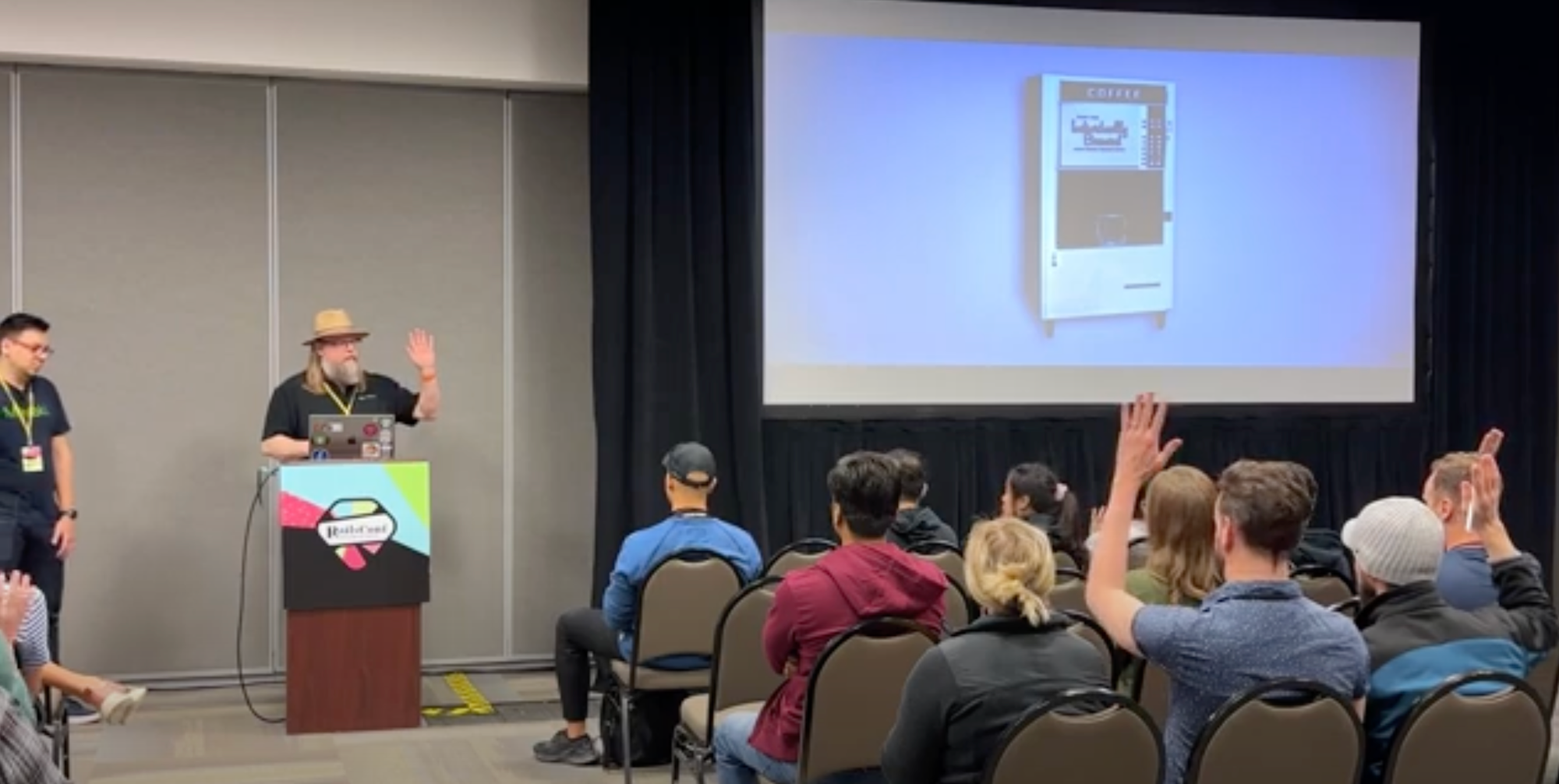 Fito von Zastrow (standing to the left) and Alan Ridlehoover (standing behind RailsConf 2023 lectern) with a picture of a mechanical coffee machine on screen. 