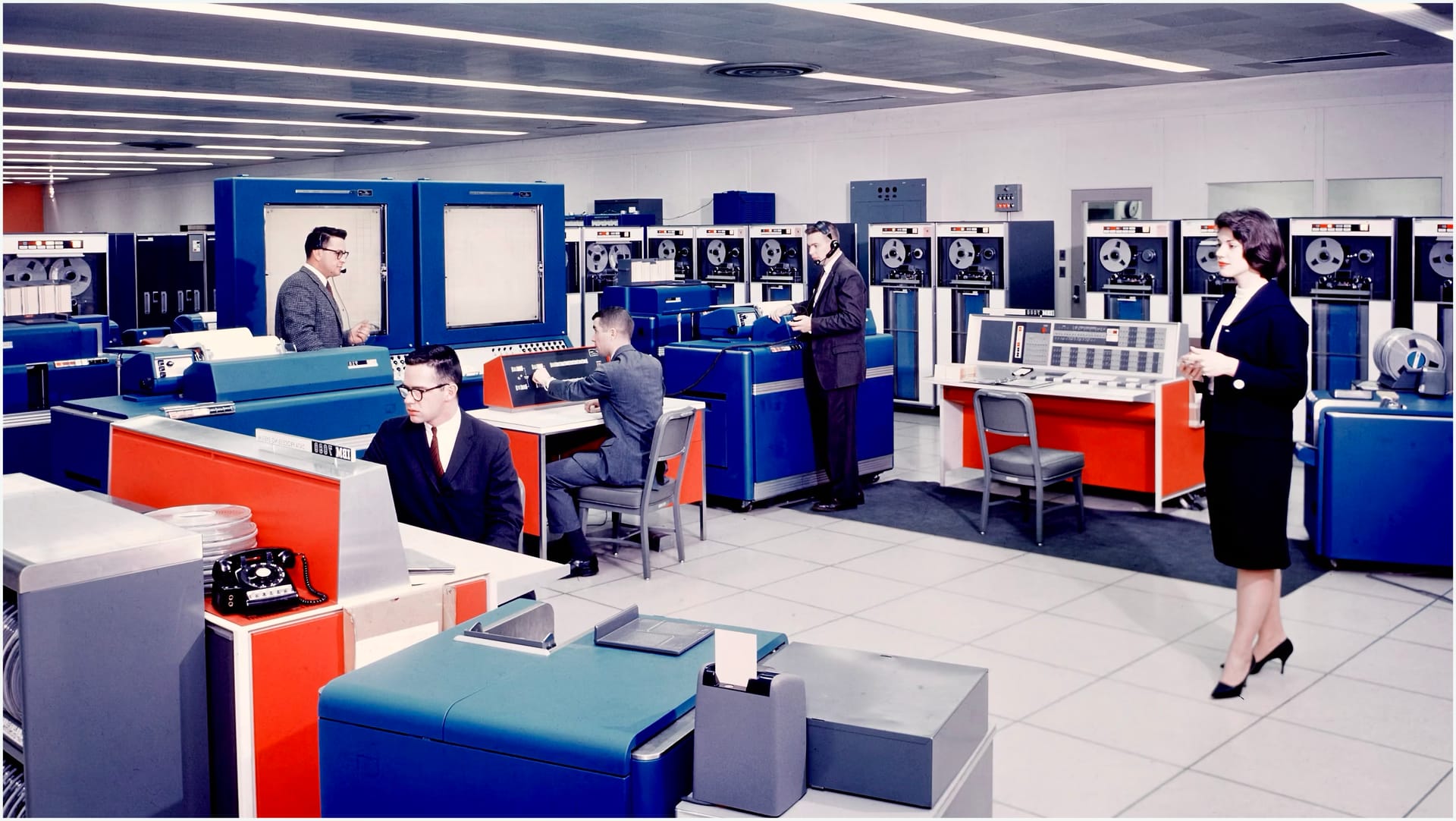 Brightly colored mainframe computer equipment with men and women in suits operating the equipment.