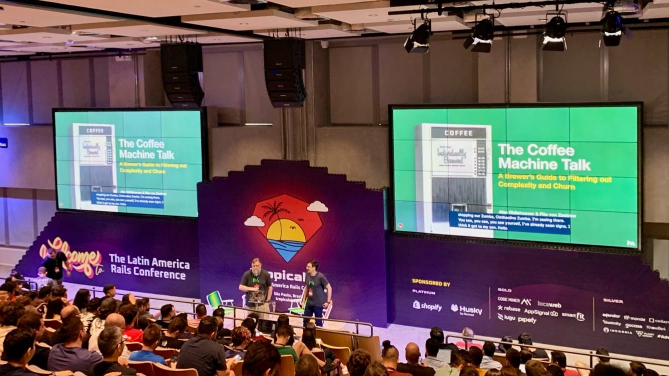 Two men speaking to a large audience in front of two giant screens with slides entitled The Coffee Machine Talk